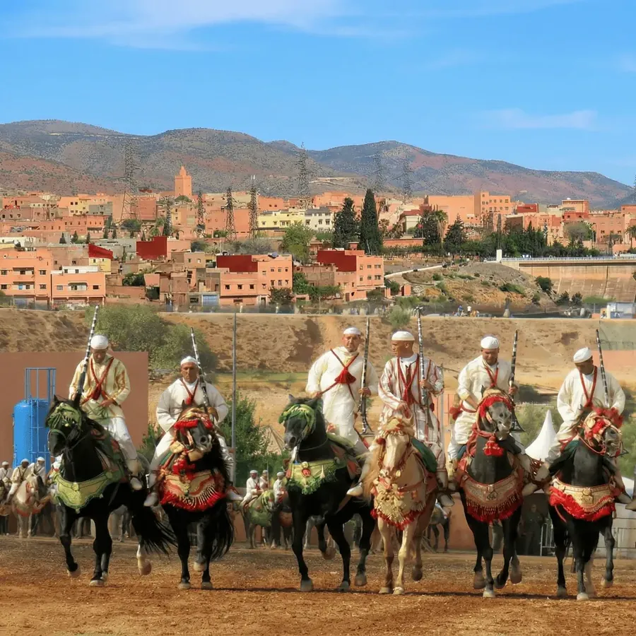 Tbourida horsemen in white robes lined up with raised muskets in front of Afourar and the Atlas foothills